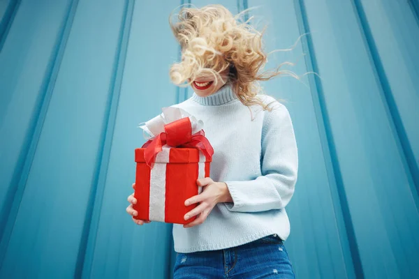 Heureuse femme blonde en tenant boîte cadeau et en regardant la caméra tout en appréciant sur fond bleu. — Photo