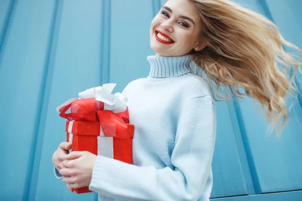 Heureuse femme blonde en tenant boîte cadeau et en regardant la caméra tout en appréciant sur fond bleu. — Photo