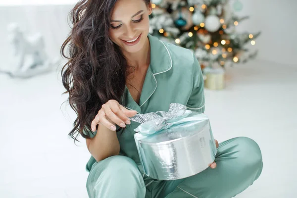 Beautiful sexy young woman sitting next to a Christmas tree holding a present present. Christmas photo. — Stock Photo, Image