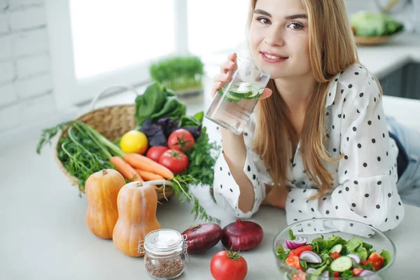 En ung kvinna dricker vatten nära bordet med frukt och grönsaker i köket. — Stockfoto