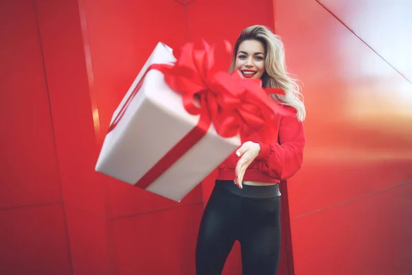 Boldog szőke nő kezében ajándék doboz és nézi a kamera, miközben élvezi a piros háttér. — Stock Fotó