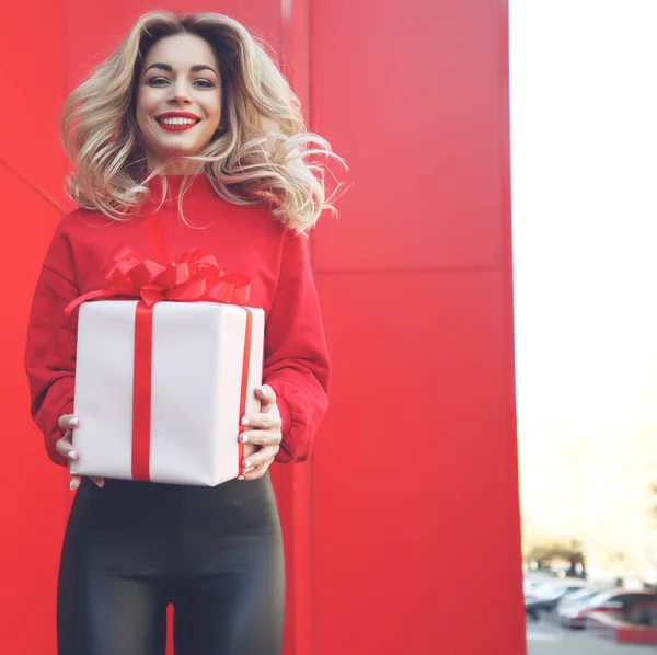 Boldog szőke nő kezében ajándék doboz és nézi a kamera, miközben élvezi a piros háttér. — Stock Fotó