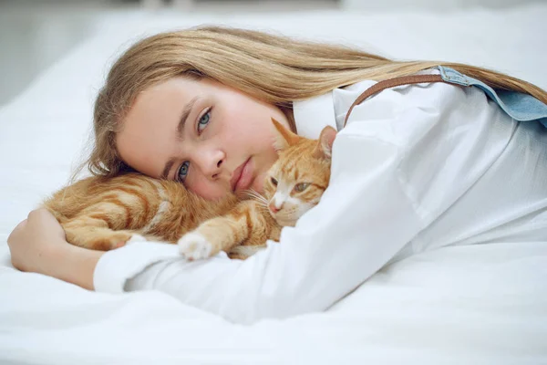 La chica está descansando en la cama con su gatito jengibre. Un niño con un animal. — Foto de Stock