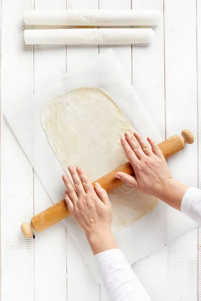 Woman hands spreading a very thin strudel dough with a rolling pin on a sheet of kitchen paper with cornstarch to prevent sticking