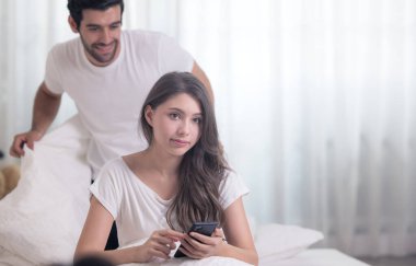 Yatakta genç bir çift, mutlu gülümseyen bir çift battaniye ve cep telefonuyla yatıyorlar.