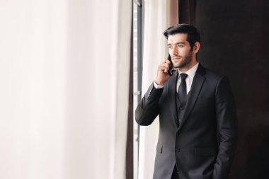 Başarılı genç işadamı, pencerelerin yanında cep telefonuyla konuşuyor.