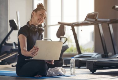 Spor giyim sektöründe sportif bir kadın dambıl, protein içeceği ya da bir şişe suyla yerde oturuyor ve evinde dizüstü bilgisayar kullanıyor. Spor ve eğlence konsepti.