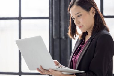 Pencerelerin yanında duran ve modern ofiste dizüstü bilgisayar kullanan iş kadını