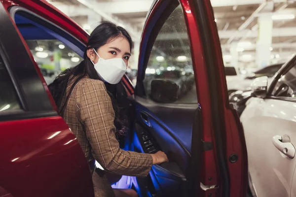 Asian woman using a surgical mask open a car door while looking at the camera at car-park background. Safety and shopping concept.