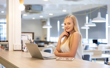 Beyaz gömlekli güzel bir kadın iş yerinde otururken dizüstü bilgisayarda çalışıyor ve gülümsüyor. . 