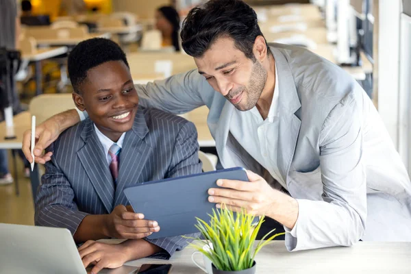 Using a computer tablet, focused black and white partners debate a project. Two multiracial male business associates