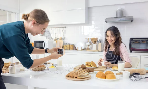 Beautiful woman pastry chef kneading bread dough on the worktop, as another female snaps a snapshot of her doing so.