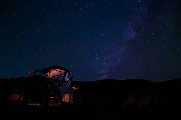 A woman sits in a farmer\'s automobile, gazing at the stars in the night sky, with the Milky Way in the background.