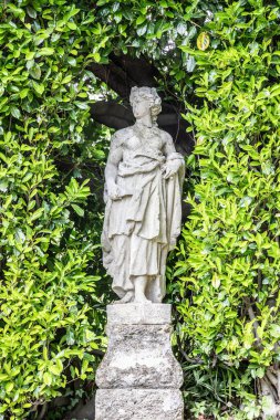 Klasik bir kadın figürünün antik taş heykeli, tarihi bir bahçedeki yemyeşil yaprakların arasında duruyor, eskimiş detayları ve sakin atmosferi gösteriyor..