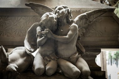 Mermer bir heykelin yakın çekimi, bir meleğin bir çocuğu nazikçe kucakladığını gösteriyor, sevgiyi, korumayı, masumiyeti ve maneviyatı ayrıntılı sanatsal ustalıkla simgeliyor..