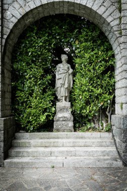 Klasik bir kadın figürünün antik taş heykeli, tarihi bir bahçedeki yemyeşil yaprakların arasında duruyor, eskimiş detayları ve sakin atmosferi gösteriyor..