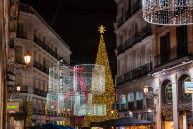 MADRID, SPAIN - 4 HAZİRAN 2026: Puerta del Sol 'da yükselen bir Noel ağacı Calle del Carmen' in sonunda parıldıyor, Madrid 'in merkezi atardamarlarından biri, güzel çerçevelenmiş şenlik ışıkları ve zarif cepheleriyle.