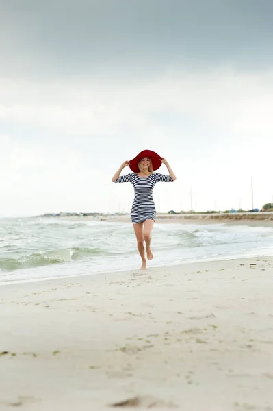 美しいですセクシーブロンド女の子でストライプドレスと赤い帽子ランに沿ってザビーチ — ストック写真