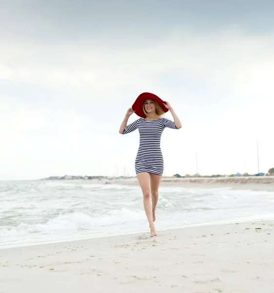 美しいですセクシーブロンド女の子でストライプドレスと赤い帽子ランに沿ってザビーチ — ストック写真