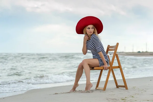 美しいですセクシーブロンド女の子でストライプドレスと赤い帽子座っています上の木の椅子オンザビーチ — ストック写真
