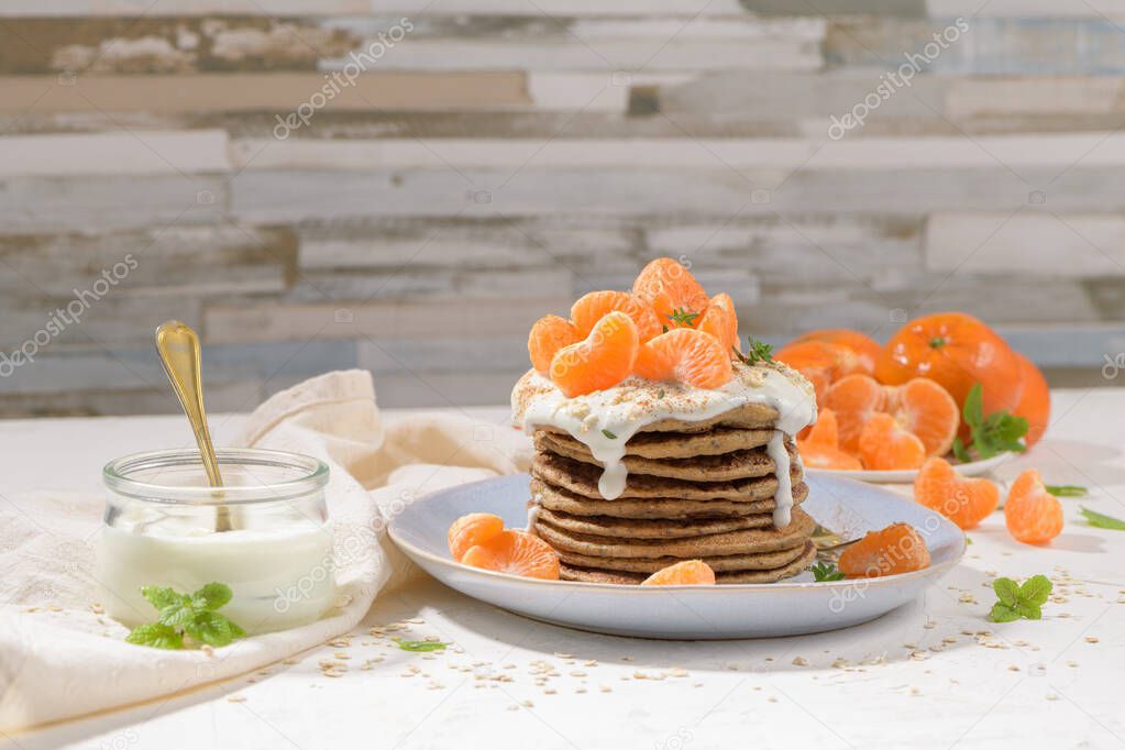 Pancakes with yogurt and tangerines on a light modern kitchen counter top with oat, thyme and mint leaves.