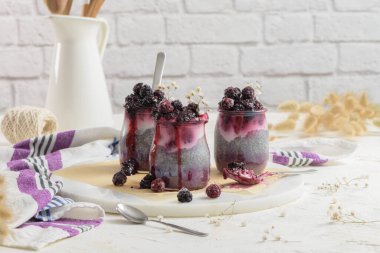 Böğürtlenli Chia pudingi, beyaz bir masada cam kavanozlarda üç porsiyon.