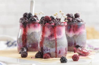 Böğürtlenli Chia pudingi, beyaz bir masada cam kavanozlarda üç porsiyon.