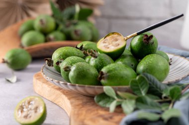 Küçük siyah bir kaşıkla beyaz flüt tabağına yerleştirilmiş taze yeşil fayjoa meyveleriyle dolu alçak açılı bir hayat. Meyvelerden biri ikiye bölünmüş, iç kısmı sararmış, iç kısmı ise su damlacıklarıyla kaplı. Kırsal bir orman.