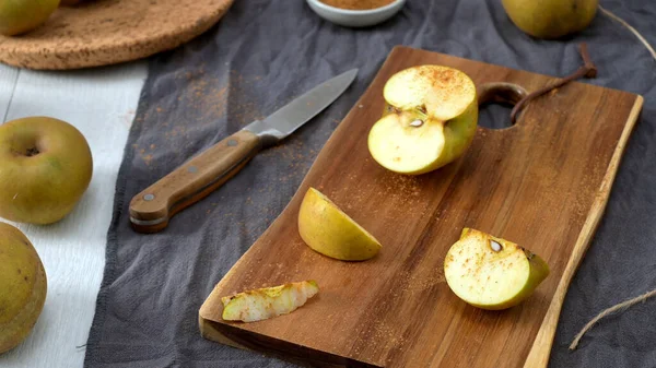 Birkaç altın elma, ahşap bir masa üzerine saçılmış kumaş ve mantar işlemeli kumaş ile. Bıçak ve kesme tahtası dilimlenmiş, tarçın tozlu bir elma sıcak, ev yapımı bir yemek için yakın bir hazırlık olduğunu gösteriyor..
