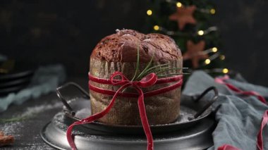 Geleneksel bir İtalyan Panettone pastasının, kadife kırmızı kurdele ve biberiye filizi ile bağlanmış, koyu metal servis standında dinlenen, kırsal ve yakın plan bir çekimi..