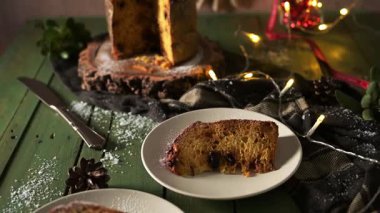 Tek bir dilim nemli panettone beyaz bir tabakta duruyor, güzel çerçevelenmiş, sicim ışıkları gibi bayram dekorasyonları ve koyu yeşil ahşap bir masa üzerindeki ebedi yeşil yapraklar gibi..