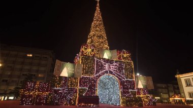 ESPINHO, PORTUGAL - 6 Aralık 2025: Altın ışıklarla aydınlatılmış devasa, konik bir Noel ağacı, istiflenmiş hediye ve kurdeleleri andıran kırmızı ve beyaz ışıklı kutulardan inşa edilmiş anıtsal bir kemer üzerinde duruyor..
