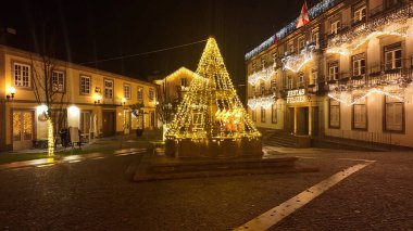 FEIRA, PORTUGAL - 5 Aralık 2025: Taştan bir çeşmeyi kaplayan sıcak, altın telli ışıklardan yaratılan büyük bir Noel ağacı şeklindeki bir şehir meydanının yakın, düşük açılı görüntüsü.