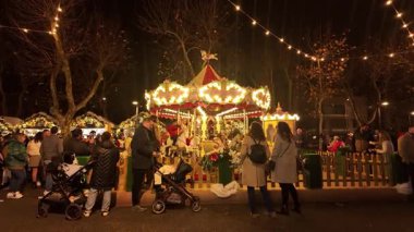VILA DO CONDE, PORTUGAL - 7 Aralık 2025: Canlı bir kalabalık, gece açık bir Noel pazarında kırmızı ve beyaz çizgili bir çatısı ve parıltılı altın ışıkları olan klasik, aydınlatılmış bir atlıkarınca etrafında toplanır.