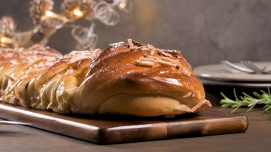 Dilimlenmiş bademler ve toz şekerle kaplanmış taze pişmiş, parlak örülmüş tatlı ekmeğin (Noel çelengi ya da Babka gibi) yakın plan, düşük açılı bir çekimi. Sıcak, davetkar sahne, karanlık bir tahta kesme tahtasının üzerinde bulanık bir şekilde geçiyor.