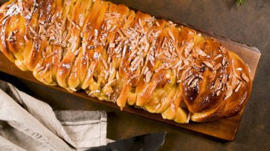 Yakından, yüksek açılı, koyu renkli bir yemek fotoğrafı taze pişmiş, parlak, örülmüş tatlı bir ekmeği gösteriyor. Üzerine dilimlenmiş bademler serpiştirilmiş ve ahşap bir kalas üzerinde sunuluyor. Aydınlatma dramatik ve sade, altın-kahverengi cru vurgulanıyor