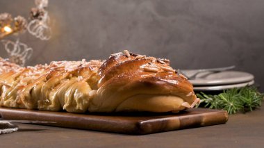 Dilimlenmiş badem ve pudra şekerle kaplanmış, neşeli, loş bir ortamda koyu bir ahşap kesme tahtasında dinlenen, altın-kahverengi, örülmüş tatlı bir ekmeğin (Vnoka veya benzeri) yakın açılı bir çekimi. Yumuşak, sıcak, aydınlık.