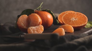 Güzel, karanlık ve karamsar bir natürmort taze, yeşil yapraklı mandalinaların yanı sıra soyulmuş segmentler ve yarım kesilmiş meyveler, koyu bir ahşap tahtada dinleniyor ve örtülü kumaş, gösterişsiz ışıklandırmayla vurgulanıyor..