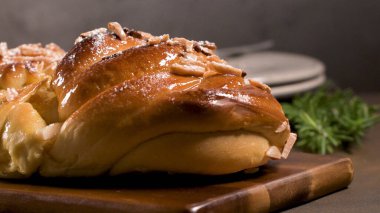 Yakın plan, düşük açılı bir çekim, yüksek parlaklıkta, üzerinde kızartılmış badem ve toz şekerle kaplı, ahşap bir kesme tahtasının üzerinde sıcak, karanlık bir ortamda dinlenen bir parça tatlı ekmek yakalar. Yumuşak, karamsar stüdyo müziği.