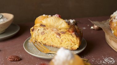 Bayramlık meyveli kekin bir kesitinde fındık ve kuru üzümlerle dolu yoğun bir dokuya yer veriliyor, üstüne de jöleli ananas halkası ve pudra şekeri. Sıcak stüdyo ışıkları kırsal havayı geliştirir..