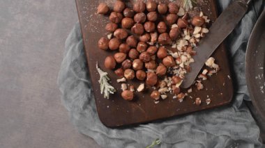 Bütün ve doğranmış fındıklar eski bir bıçakla birlikte koyu bir ahşap tahtaya saçılmış. Kompozisyonda kırsal bir mutfak hissi için karamsar ışıklandırma ve yumuşak kumaş dokusu yer alıyor..