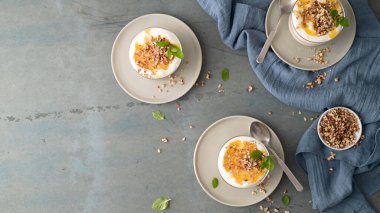 Üzerinde altın bal, doğranmış fındık ve taze nane bulunan kremalı yoğurt kompozisyonu. Yumuşak doğal ışıklandırma, desenli mavi kumaş ve kırsal gri taş arka planı güçlendirir..