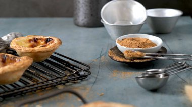 Taze pişmiş Portekiz yumurtalı turtaları bir kase tarçınla birlikte soğutma rafında otururlar. Köy sahnesinde karamsar ışıklandırma, desenli mavi yüzeyler ve eski metal ölçüm kaşıkları yer almaktadır..