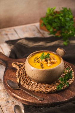 Sıcak bir kase tereyağlı kabak çorbası ahşap bir tahtanın üzerinde kırsal seramik bir tencerede servis edilir. Sahnede toprak dokular, yumuşak doğal ışıklandırma ve taze maydanoz süsleri yer alıyor..