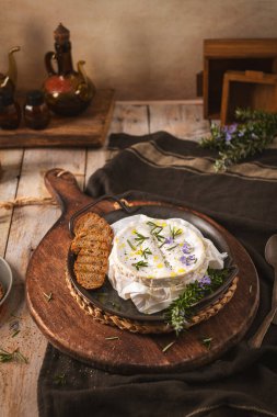 Erimiş Camembert peynirinin bir gurme tekerleği, koyu metal bir tavada sunulan taze bitkiler ve yağla kaplıdır. Dikey kompozisyonda sıcak, sinematik ışıklandırma ve sıcacık bir çiftlik evi estetiği yer alıyor..