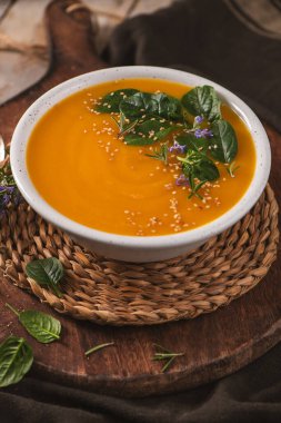 Bir kase canlı portakallı tereyağlı kabak çorbası ya da kabak çorbası rustik ahşap bir tahtanın üzerinde servis edilir. Sahnede, püre ve otların pürüzsüz dokusunu vurgulayan ılık doğal ışıklandırma yer alıyor..