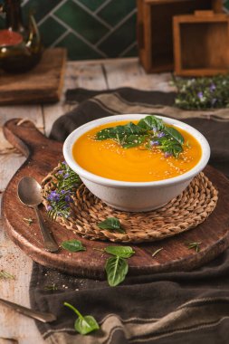 Canlı bir portakallı sebze püresi taze ıspanak, susam tohumu ve biberiye ile süslenir. Kaba gıda fotoğrafçılığında sıcak ışıklandırma ve ahşap elementler yer alıyor..