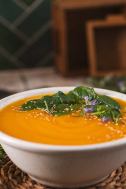 Kırsal seramik bir kasede canlı bir tereyağlı kabak ya da kabak çorbası servis edilir. Tabakta taze yeşiller, mor çiçekler ve sıcak, yumuşak doğal ışıklandırma altındaki tohumlar yer alıyor..