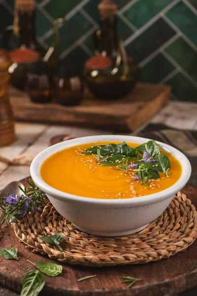 Canlı bir portakal kabağı çorbası sıcak bir mutfak ortamında dokuma hasırının üzerinde duruyor. Yumuşak dokusu kızarmış susam tohumları ve yenilebilir mor çiçeklerle vurgulanır..