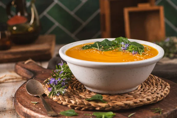 Ispanak, biberiye ve susam tohumlarıyla süslenmiş canlı turuncu sebze püresi. Kase, örülmüş bir bardak altlığı ve ahşap bir tahtanın üzerinde, sıcak, sığ alan derinliğinde ışıklandırmayla yakalanmış..